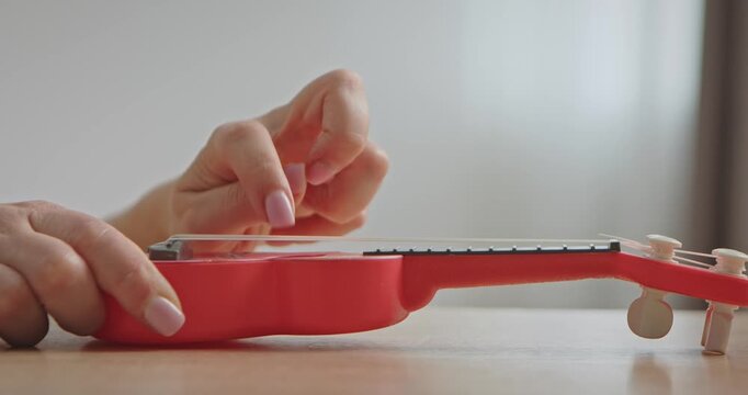 Close up of a hand playing the strings of a red toy ukulele on a table. Stringed musical instrument on a wooden table in a bright room.