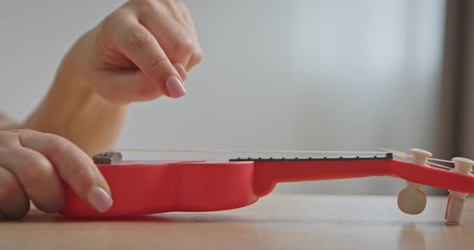 Close up of a hand playing the strings of a red toy ukulele on a table. Stringed musical instrument on a wooden table in a bright room.