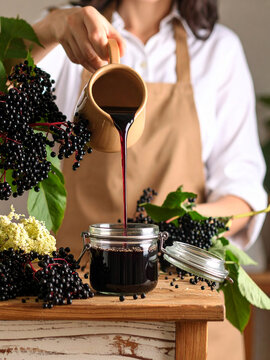 A person in an apron is pouring dark liquid from a pitcher into a jar. Elderberries are arranged around the jar and pitcher on a wooden surface.