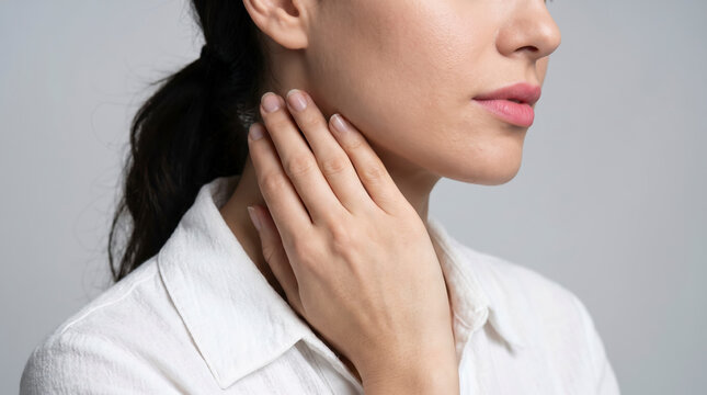 Young woman experiencing neck pain while touching her cervical spine with hand on light gray background in close up studio shot for medical healthcare concept