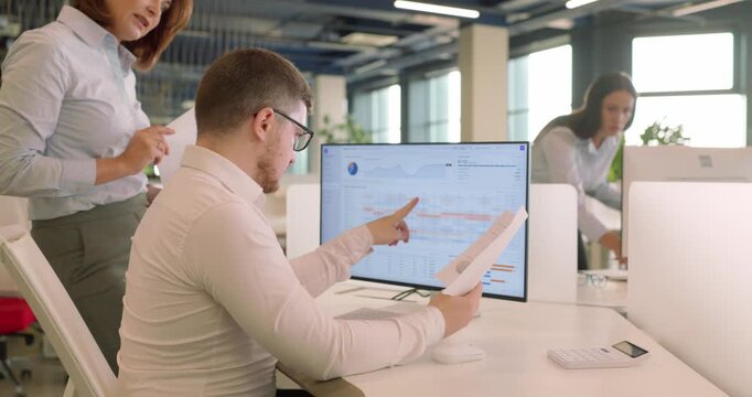 Analyst and manager check budget report at computer. A man compares a paper printout with financial data on the screen while a colleague advises. Teamwork in budget planning and analysis.