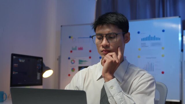 Pensive young programmer analyzing complex data on laptop computer during overtime work shift in tech office