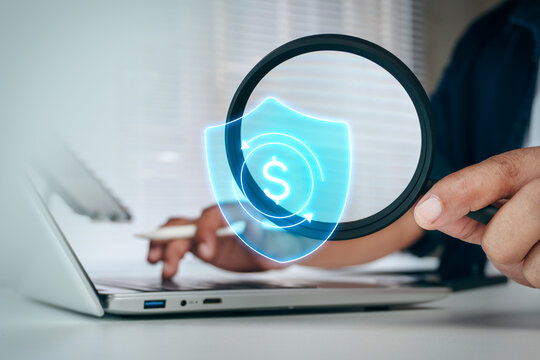 Business professional holding magnifying glass to inspect digital dollar shield icon overlay on laptop screen, concept of financial protection, secure banking, and payment fraud prevention.