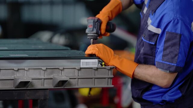 Close-up of gloved hands in a blue work shirt driving a screw into the edge of an EV battery module with a cordless drill, steadying the casing with the other hand