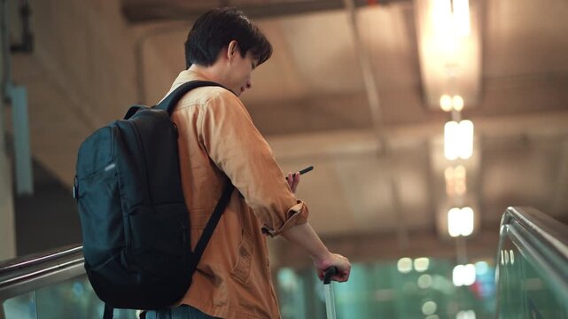 Young Asian man with backpack checking smartphone while riding up an escalator at modern airport terminal, male traveler pulling wheeled suitcase during journey, air travel.