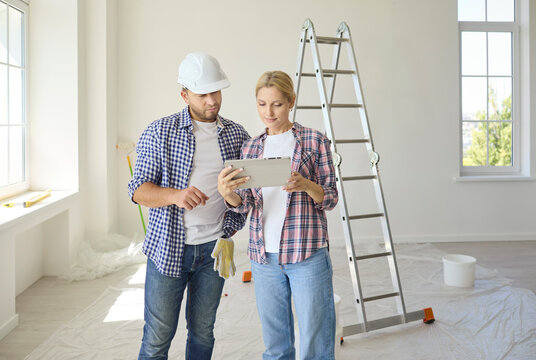 Young couple reads renovation estimate on tablet. Husband in hard hat and wife calculate cost of home repair. The room has white walls and big windows. Home improvement and teamwork concept.