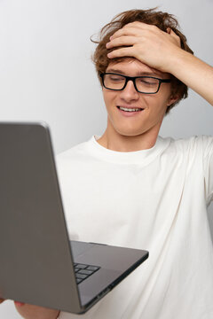 Amused young man with glasses laughing while holding laptop, hand on head. Relatable tech mistake, lighthearted computer problem, friendly user experience concept.