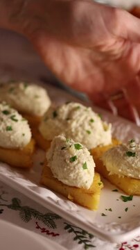 Extreme close-up of hands placing a plate of grilled polenta squares topped with traditional Italian whipped salt cod cream, known as baccal&agrave; mantecato. A festive Italian Christmas appetizer featuring