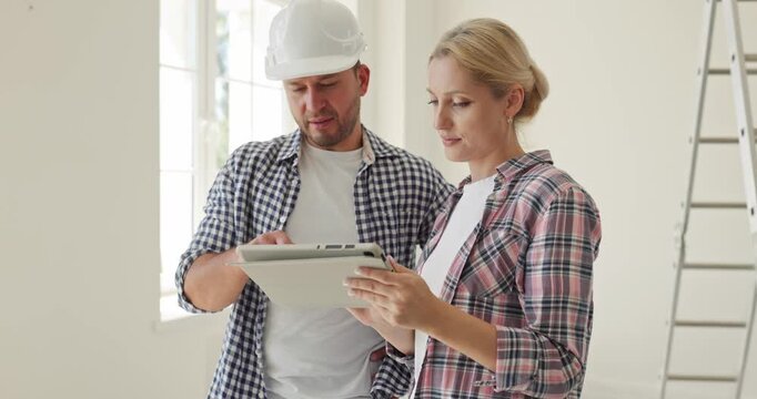 Couple planning apartment renovation using tablet. A contractor in helmet reviews homeimprovement design details with homeowners beside a ladder. Practical home improvement planning concept.