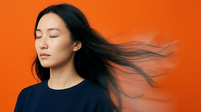 Serene Asian woman with eyes closed, blowing long black hair in wind motion on vibrant orange background, beauty studio portrait