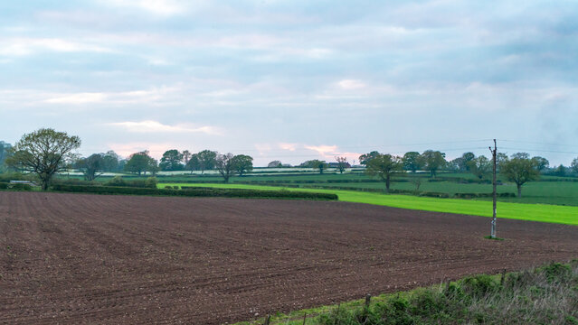 A rural agricultural landscape with a plowed field in the foreground and green meadows and forest strips in the background