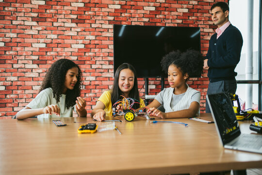 Three schoolgirls build a small wheeled robot with an adult instructor in a classroom