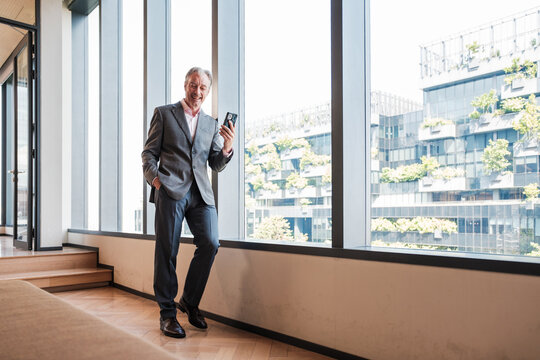 Smiling senior businessman checks his smartphone beside windows in a modern office