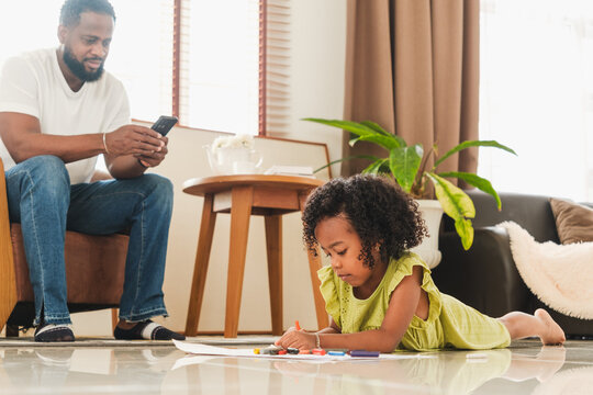 A young girl draws on the living room floor while her father looks at his phone nearby