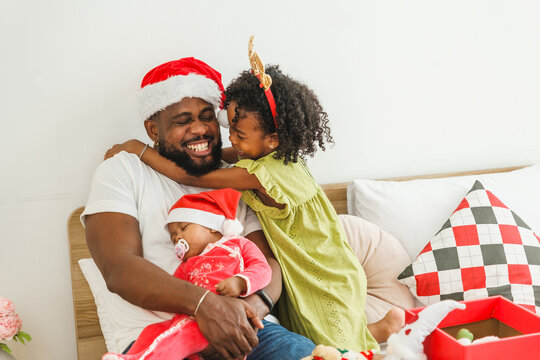 Father in a Santa hat embraces his daughter while holding their sleeping baby at home for Christmas