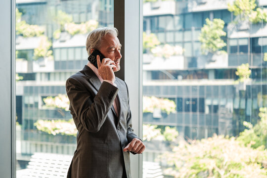 Mature businessman talks on a smartphone beside a floor to ceiling office window