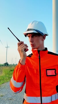 Wind farm technician in high visibility workwear checks his sleeve and speaks into a radio beside turbines
