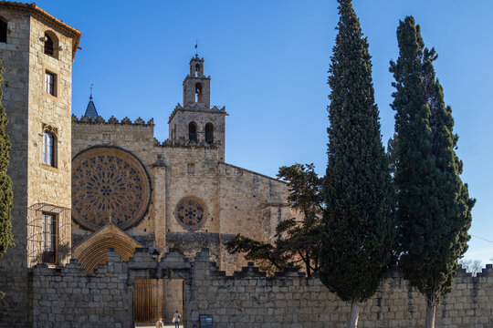 Rose window and pointed Gothic portal on the facade of the Monestir de Sant Cugat with tall cypress trees and a crenellated stone wall under a blue sky