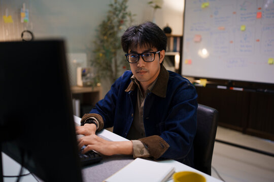 Asian male software developer looking at computer monitor screen working late night home office focused programmer technology engineer professional
