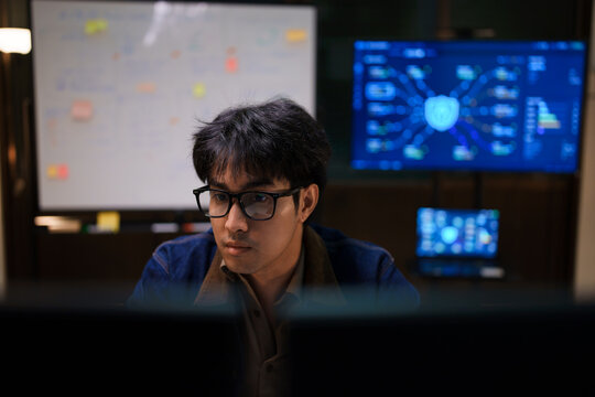 Asian male IT developer looking concentrated at computer screen working late night on cybersecurity network and data protection system in office