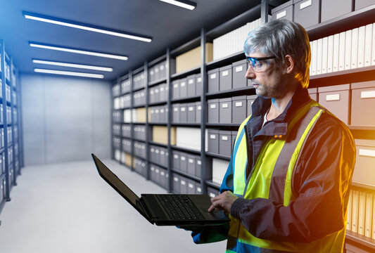 Document archive management, technician uses a laptop to inspect and organize stored files, records and boxes in a secure industrial archive room.