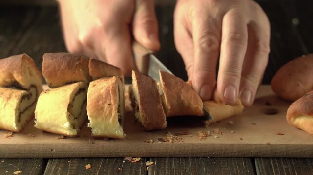 Hands slicing a rolled loaf of bread into pieces on a wooden cutting board, showing fresh pastry texture and crumbs in a warmly lit kitchen setting