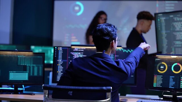 Black developer points at lines of code on a big screen while an Asian man in a suit at the front desk raises his hand to ask a question during a late night AI lab briefing
