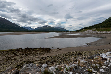 Wide view of arctic fjord coastline featuring tidal sandbars, rocky foreground, and distant snow capped peaks in Breivikeidet, Norway © LAURA