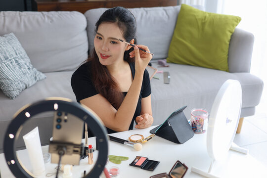 Asian woman filming beauty vlog tutorial with ring light, reviewing cosmetics and makeup concept.