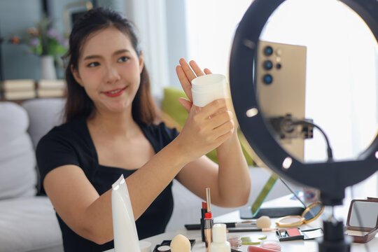Asian woman filming beauty vlog tutorial with ring light, reviewing cosmetics cream moisturizer or lotion.
