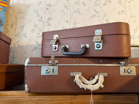 two stacked vintage brown leather suitcases with metal clasps and rope handle against patterned floral wallpaper, nostalgic travel luggage in retro interior