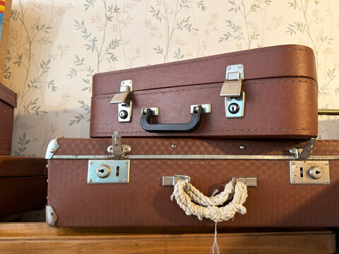 stack of two old brown travel suitcases with metal locks and rope handle against faded floral wallpaper, vintage luggage and nostalgic travel concept