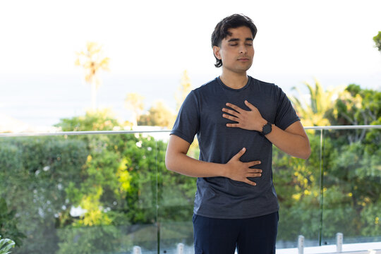 Indian man standing on glass balcony wearing gray T-shirt and watch, hands on chest and abdomen