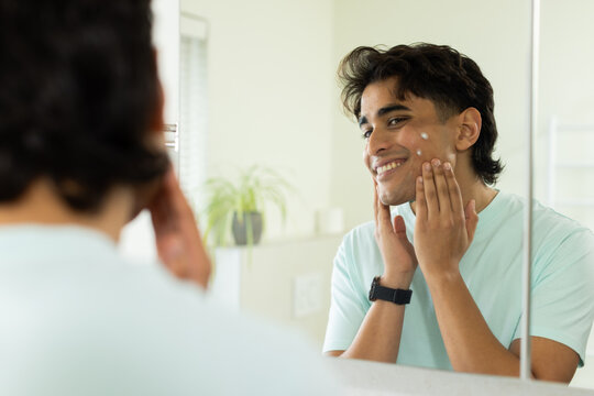 African American man in mint tee applying facial cream at bathroom mirror, showing dark watch