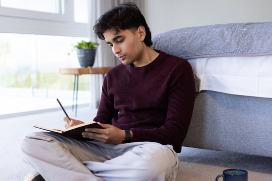 African American man in maroon sweater sitting on bedroom floor writing in notebook with pen