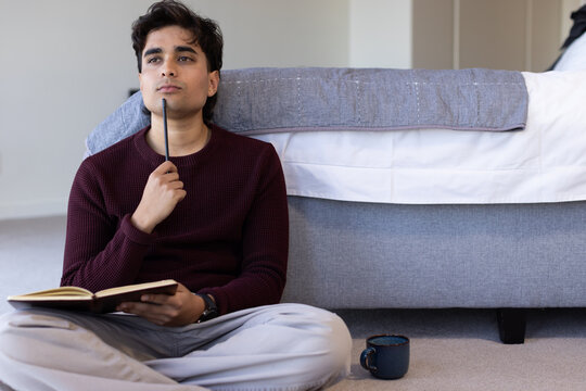 Man sitting on carpeted bedroom floor in maroon sweater holding notebook and pen