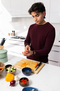 Indian man peeling carrot at kitchen island, wearing burgundy sweater beside peeler on wooden board