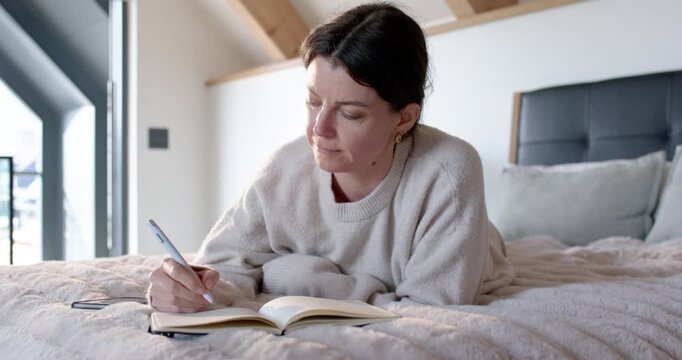 Woman in thirties writing notes in notebook on bed in loft bedroom, checking smartphone after alert