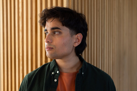 Asian man mid-20s standing against vertical-slat wall wearing green button-up over rust tee