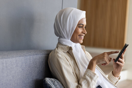 Adult woman in white hijab, striped shirt smiling, tapping black smartphone on gray sofa