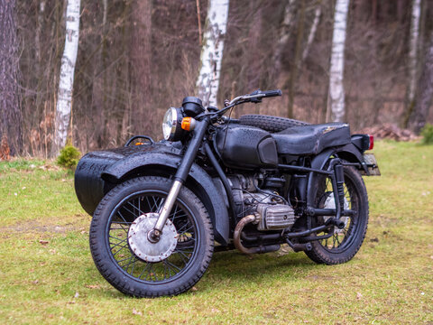 A vintage matte black motorcycle equipped with a sidecar stands parked on a grassy lawn near birch trees outdoors.