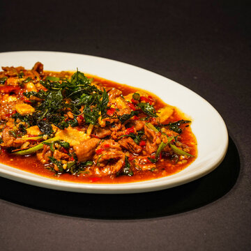 Thai holy basil chicken stir-fried with red chilies and savory sauce, topped with crisp basil leaves and served on a white plate against a plain dark background, showcasing a spicy Thai-inspired meal.