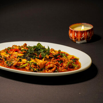 Thai holy basil chicken stir-fried with red chilies and savory sauce, topped with crisp basil leaves and served on a white plate against a plain dark background, showcasing a spicy Thai-inspired meal.