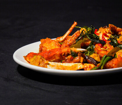 Crab pieces and claws coated in golden yellow curry sauce with vegetables, peppers, and herbs, served on a white plate against a dark background, showcasing a savory seafood dish for restaurant menus.