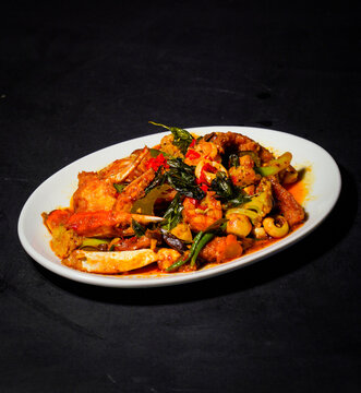 Crab pieces and claws coated in golden yellow curry sauce with vegetables, peppers, and herbs, served on a white plate against a dark background, showcasing a savory seafood dish for restaurant menus.