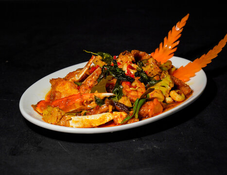 Crab pieces and claws coated in golden yellow curry sauce with vegetables, peppers, and herbs, served on a white plate against a dark background, showcasing a savory seafood dish for restaurant menus.