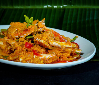 Crab pieces and claws coated in golden yellow curry sauce with vegetables, peppers, and herbs, served on a white plate against a dark background, showcasing a savory seafood dish for restaurant menus.