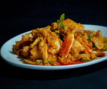 Crab pieces and claws coated in golden yellow curry sauce with vegetables, peppers, and herbs, served on a white plate against a dark background, showcasing a savory seafood dish for restaurant menus.