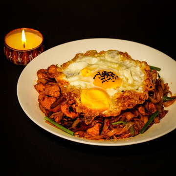 Thai drunken noodles stir-fried with meat, vegetables, and green onions, topped with a fried egg and chili flakes, served in a white bowl against a dark background, a savory restaurant dish for menus.