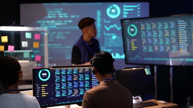 Black engineer in a blue vest presents in front of a giant code wall screen, writing with a stylus on a tablet while two seated teammates in headsets watch from their desks